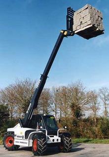 Ładowarka teleskopowa Bobcat T40140 podnosi paletę z kostką brukową na tle błękitnego nieba i drzew bez liści.